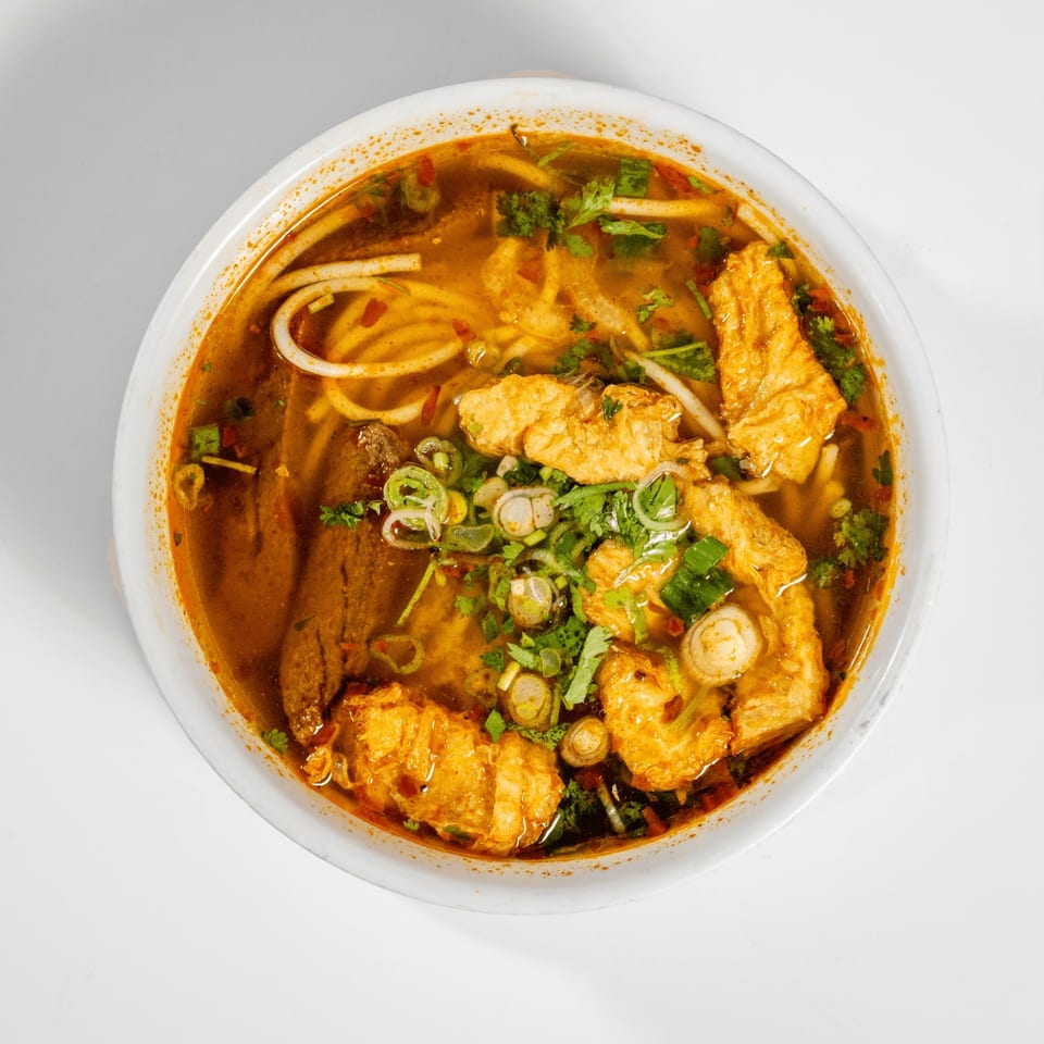 Overhead view of a bowl of spicy Vietnamese noodle soup with fried tofu and herbs.