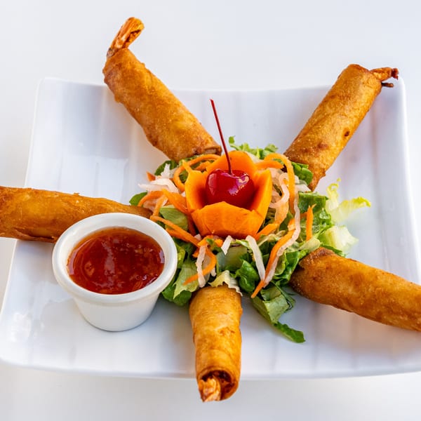 Five crispy shrimp spring rolls arranged around a fresh salad with a cherry garnish and dipping sauce.