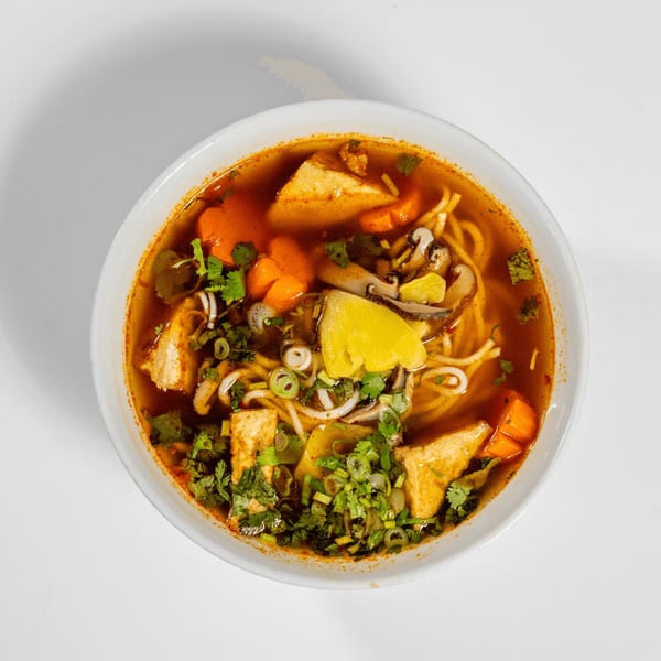 A bowl of spicy noodle soup with tofu, carrots, mushrooms, pineapple, and herbs.