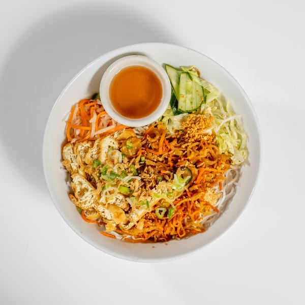 A white bowl of vermicelli noodles with tofu, shredded carrots, cabbage, and a side of sauce.