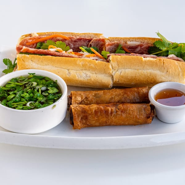 Banh mi sandwich with spring rolls and two dipping sauces on a white plate.