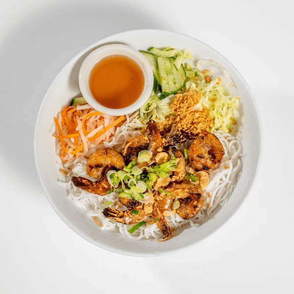 A white bowl of vermicelli noodles with grilled shrimp, shredded vegetables, and a side of sauce.