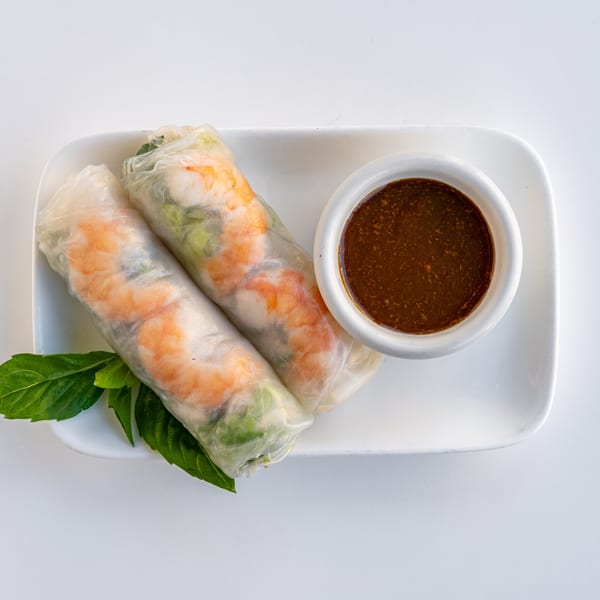 Two fresh shrimp spring rolls with basil leaves and a side of dark dipping sauce on a white plate.