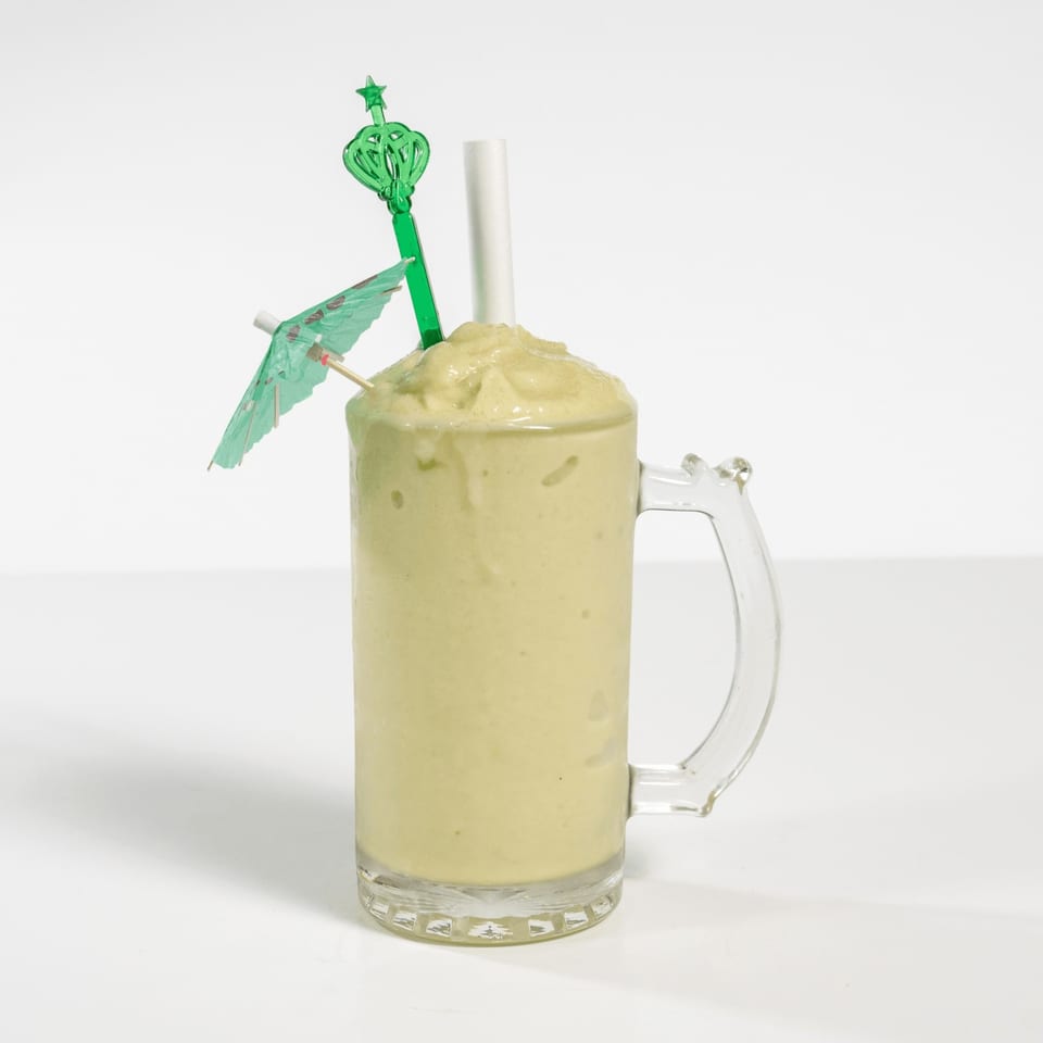A light green smoothie in a glass mug with a green umbrella and star-shaped stirrer.