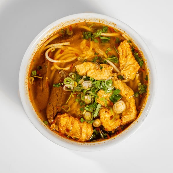 Overhead view of a bowl of spicy Vietnamese noodle soup with fried tofu and herbs.