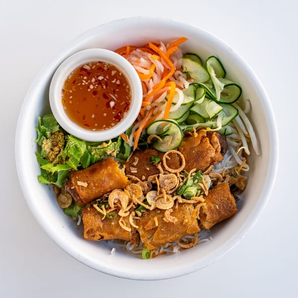A white bowl filled with Vietnamese vermicelli noodles, crispy spring rolls, fresh vegetables, and a side of dipping sauce.